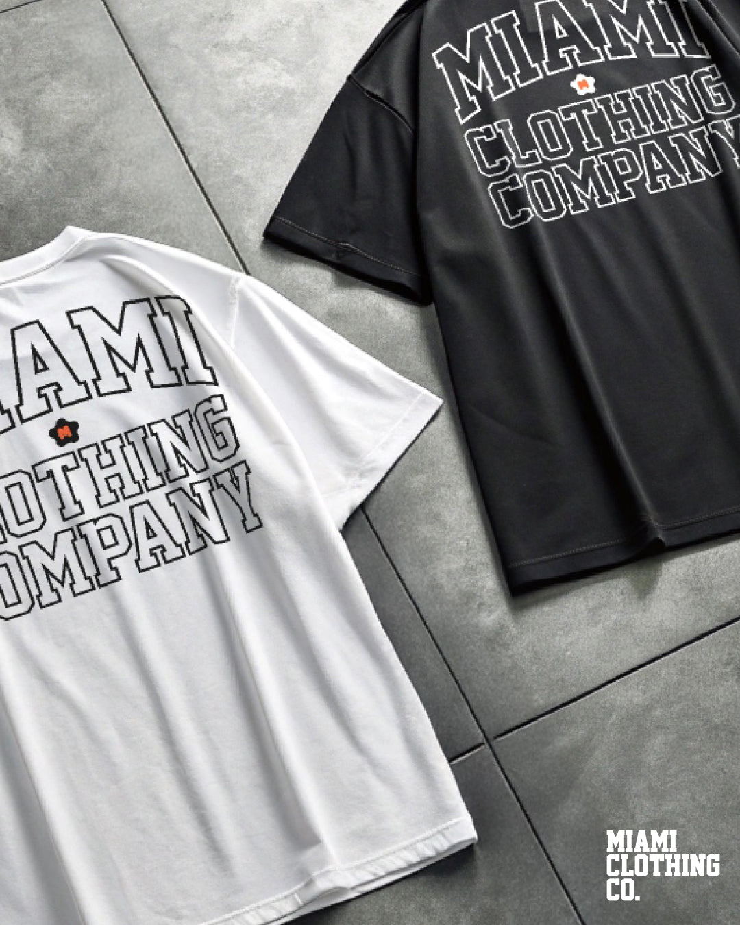 Flat lay of black and white Miami Clothing Company Signature Tees showing the arched back logo design on concrete flooring.